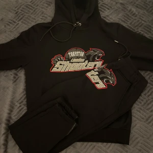Svart hoodie från Trapstar - Säljer en svart hoodie från Trapstar med ett stort broderat motiv på framsidan. Hoodien har en justerbar dragsko i huvan och ribbade muddar vid ärmslut och nederkant. Perfekt för en avslappnad stil. BARA HOODIE!!!! 