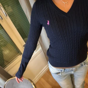 Mörkblå tröja från Ralph Lauren - Säljer en stilren mörkblå kabelstickad tröja från Ralph Lauren med en rosa broderad logotyp på bröstet. Tröjan har en v-ringad hals och långa ärmar, perfekt för en klassisk look. Passar bra till jeans eller kjol för en avslappnad men elegant stil.