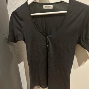 Mörkgrå kortärmad topp från Acne Jeans stl S - Säljer en stilren svart topp från Acne Jeans med korta ärmar och knappar framtill. Toppen har en v-ringad design och är perfekt för en avslappnad look. Storlek S 100% bomull.