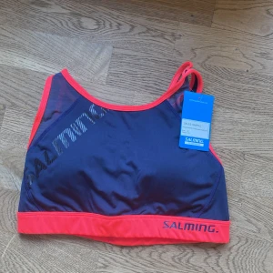 Blå och röd sport-bh från Salming - Säljer en sportig och bekväm blå sport-bh från Salming med röda detaljer. Den har en snygg korsad ryggdesign och elastisk kant för extra stöd. Perfekt för träning och aktivitet.