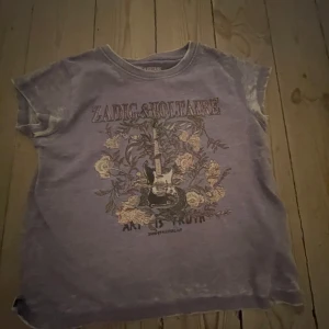 Lila t-shirt från Zadig & Voltaire - Säljer en cool lila t-shirt från Zadig & Voltaire med ett rockigt tryck av en gitarr och blommor. T-shirten har korta ärmar och en avslappnad passform. Perfekt för en avslappnad stil till sommaren, köpt för 500 säljer för 200