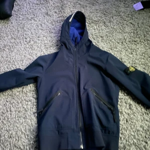 Mörkblå jacka med huva från Stone Island - Säljer en mörkblå jacka med huva från Stone Island. Jackan har dragkedja framtill och två fickor med dragkedjor. Den har en ikonisk logga på ärmen och är perfekt för kyligare dagar.