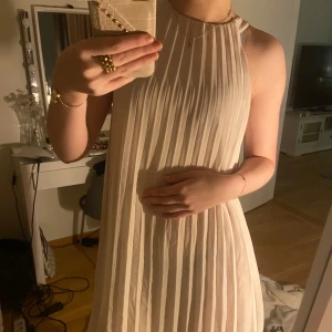 Beige ärmlös klänning - Säljer en elegant beige ärmlös klänning med plisserad design. Perfekt för en stilren look. Klänningen har en luftig passform och är tillverkad i ett lätt material.