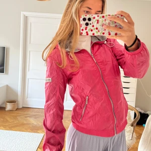 Rosa vindjacka  - Säljer en snygg rosa vindjacka  med dragkedja och två fickor framtill. Jackan har en normal passform och är perfekt för vår och sommar. Den har en diskret logga på ärmen och en grå insida vid kragen.