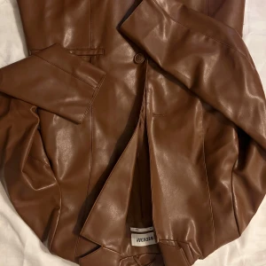 Brun skinnjacka/kavaj från Weekday - Snygg brun skinnjacka/kavaj  från Weekday i en klassisk design. Jackan har en stilren look med knappar framtill och långa ärmar. 