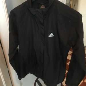 Svart vindjacka från Adidas stl small - Säljer en svart vindjacka från Adidas med Clima365-teknologi. Jackan har en dragkedja framtill och Adidas-loggan på bröstet. Perfekt för träning eller som en lätt jacka för blåsiga dagar.