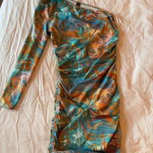 Färgglad mönstrad klänning - Säljer en färgglad klänning med ett abstrakt mönster i blått, orange och grönt. Klänningen har en one-shoulder design och långa ärmar. Perfekt för att sticka ut med sin unika stil. 🌈