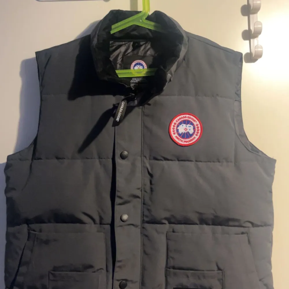 Säljer en stilren grå dunväst från Canada Goose med deras ikoniska logga på bröstet. Västen har knappar framtill och två praktiska fickor. Perfekt inför hösten och dessutom helt ny väst i storlek M. KAN KÖPAS ENDAST GENOM VINTED ELLER POST då det ej går att sälja på denna app. Och mitt vinted användarnamn är - Glass3451 . Takit.