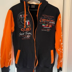 Svart och orange huvtröja från Violento - Säljer en cool svart och orange huvtröja från Violento. Tröjan har dragkedja framtill och tryck med text och grafik på bröstet. Den har långa ärmar och en bekväm huva. Perfekt för en avslappnad stil.