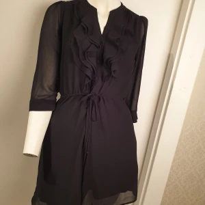 Svart klänning med volangdetaljer - Elegant svart klänning med volangdetaljer framtill och knytband i midjan. Klänningen har trekvartsärmar och en lätt transparent look som ger en sofistikerad touch. Perfekt för en stilren och chic look.