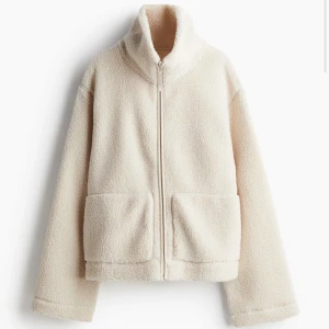 Beige teddyjacka med dragkedja - Mysig beige teddyjacka med dragkedja framtill och stora fickor. Perfekt för kyliga dagar med sin fluffiga och varma design. Jackan har en hög krage och långa ärmar för extra värme.