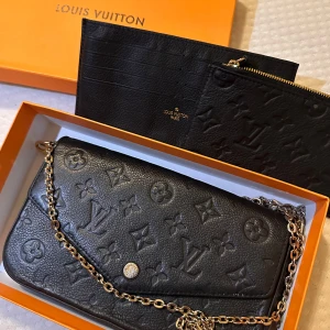 Axelremsväska  - Snygg svart axelväska med klassiskt monogrammönster. Väskan har en elegant kedjerem och du får med 2 ”plånböcker”. Väskan är som ny men har tyvärr blivit lite platt men det går att lösa när man fyller väskan
