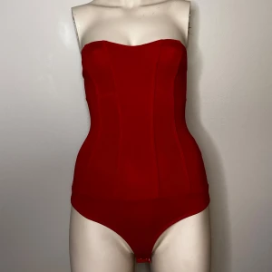 Röd bandeau body från Bershka - Röd bandeau body från Bershka i en tight passform. Den är axelbandslös och har en korsett design som framhäver figuren. Knäppen nertill.