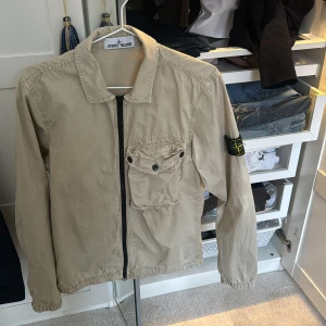 Beige jacka från Stone Island - Säljer en snygg beige jacka från Stone Island med dragkedja och en praktisk bröstficka med knapp. Jackan har en klassisk krage och det ikoniska Stone Island-märket på ärmen. Perfekt för en stilren look. Nu när våren är på ingång så passar nog denna perfekt, knappt använd skicket är 9/10. Först till kvarn då priset är redan sänkt från 1500  
