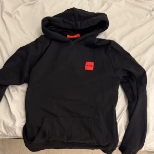 Svart hoodie från Hugo Boss - Snygg svart hoodie från Hugo Boss med en röd logotyp på bröstet. Tröjan har en klassisk design med huva och en praktisk magficka. Perfekt för en avslappnad stil. Priset kan gå ner i snabbaffär!