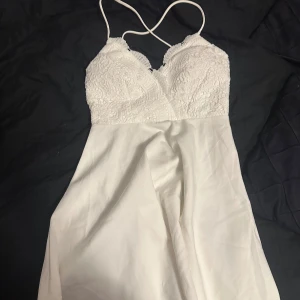 Vit klänning med spetsdetaljer - Säljer en elegant vit klänning med spetsdetaljer på överdelen och tunna axelband. Klänningen har en vacker v-ringning och är perfekt för sommarens festligheter. Den är tillverkad i ett lätt och bekvämt material som ger en fin passform.