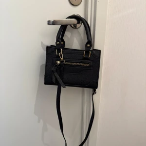 Svart väska  - En väldigt söt och fin svart väska köpt på Shein. Den har ett axelband som kan tas bort om man vill. Den är väldigt praktisk och rymmer någorlunda mycket saker. Väskan har använts några gånger, men har inga defekter och är i bra skick. Dock har den tidigare guldiga färgen börjat blekna: försvinna lite. Väskans höjd är ca 13 cm och bredd 19 cm🤍