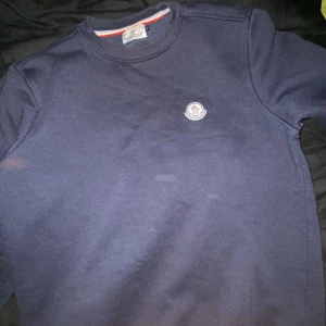 Mörkblå tröja från Moncler - Säljer en stilren mörkblå tröja från Moncler med rund halsringning och ett litet broderat Moncler-märke på bröstet. Perfekt för en avslappnad och snygg look.