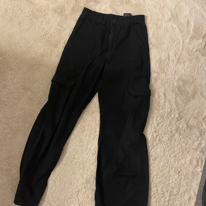 Svarta cargobyxor från H&M - Snygga svarta cargobyxor från H&M med praktiska fickor på sidorna. Byxorna har en klassisk design med knapp och dragkedja framtill. Perfekta för en avslappnad stil.