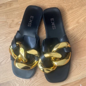 Svarta sandaler med guldkedja från SKA - Stilrena svarta sandaler från SKA med en iögonfallande guldkedja som detalj. Perfekta för att ge en touch av glamour till din outfit. Sandalerna har en öppen design och är tillverkade i syntetmaterial.
