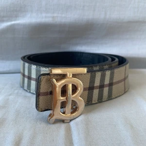 Burberry bälte - Snyggt bälte med ett klassiskt rutigt mönster i beige och bruna toner. Bältet har ett iögonfallande guldspänne format som ett 'B'. Perfekt för att ge en stilren touch till din outfit. Pris kan diskuteras vid snabb affär