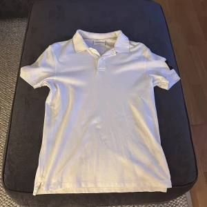 Vit pikétröja från Calvin Klein Jeans - Säljer en stilren vit pikétröja från Calvin Klein Jeans. Tröjan har korta ärmar och en klassisk krage med knappdetalj. Perfekt för en avslappnad men ändå snygg look. Märkeslogga på ärmen ger en extra touch av stil.