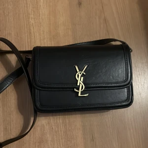 Svart axelväska från YSL - Snygg svart axelväska från YSL med ikonisk logga i guld på framsidan. Väskan har en stilren design och justerbar axelrem. Perfekt för att ge en lyxig touch till din outfit.