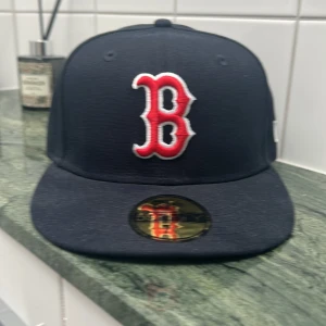Blå keps med röd broderad bokstav. Aldrig använd! - Snygg svart keps från New Era med en stor röd broderad bokstav på framsidan. Kepsen har en platt skärm och en liten logotyp på sidan. Perfekt för att ge din outfit en sportig touch.