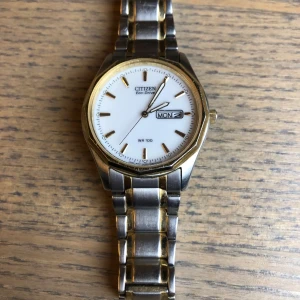 Citizen Eco-Drive klocka - Säljer en stilren Citizen Eco-Drive klocka med vit urtavla och dag/datumvisning. Klockan har en tvåfärgad länk i silver och guld samt en guldfärgad boett. Perfekt för den som gillar klassisk design.
