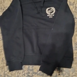 Svart hoodie med tryck - Säljer en svart hoodie med tryck på både fram- och baksidan. Framsidan har en logotyp med texten 'Rimbo IF 1911' och baksidan har flera tryck inklusive 'OKQ8 Rimbo' och 'AQUA VVS'. Hoodien har långa ärmar och en klassisk huva med snören.