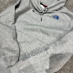 Grå hoodie från The North Face - Säljer en stilren grå hoodie från The North Face med logga i blått på bröstet och ärmen. Tröjan har en klassisk känguruficka och justerbar huva med snören. Perfekt för en relaxad stil.