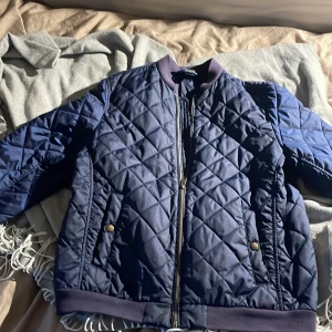 Mörkblå quiltad bomberjacka från Polo Ralph Lauren - Säljer en snygg mörkblå quiltad bomberjacka från Polo Ralph Lauren. Jackan har dragkedja och ribbade muddar vid ärmslut och nederkant. Perfekt för kyligare dagar med sin stilrena design och bekväma passform.