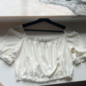 Vit blus från H&M Divided - Säljer en söt vit blus från H&M Divided med puffiga korta ärmar och elastiska kanter. Blusen har en romantisk stil med volangdetaljer vid ärmslut och nederkant. Perfekt för en somrig look!