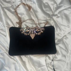 Svart kuvertväska med guldkedja - Elegant svart kuvertväska i sammet med en glittrig guldkedja och dekorativa stenar på locket. Perfekt för en kväll ute eller speciella tillfällen. Väskan har en praktisk öppning och en lyxig känsla.