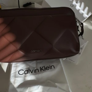 Brun Calvin Klein väska - Brun Calvin Klein väska.  Aldrig använd, prislappen är kvar. Kommer med både långa och korta band så fungerar som både handväska och axelväska.