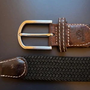 Svart flätat bälte från Billybelt - Snyggt svart flätat bälte från Billybelt med bruna skinn-detaljer och en stilren silverfärgad spänne. Perfekt för att ge en touch av stil till din outfit. Bältet har en klassisk design med kontrasterande sömmar som ger det en unik look.