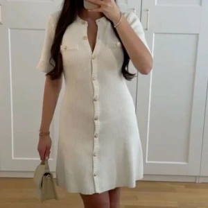 Vit stickad klänning med knappar - Snygg vit stickad klänning med korta ärmar och dekorativa knappar framtill. Klänningen har en figurnära passform och två små fickdetaljer på bröstet. Perfekt för en stilren look! Säljer denna då den aldrig använts lapp kvar storlek S.