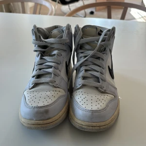 Nike sneakers i vitt - Säljer ett par vita Nike sneakers med svart swoosh. Skorna har snörning och en hög modell med vadderad krage för extra komfort. Perfekta för en sportig look.