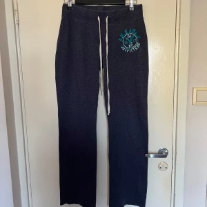 Lågmidjade mörkblå mjukisbyxor från American Eagle - Säljer ett par mörkblå mjukisbyxor från American Eagle med tryck i turkos på ena benet. Byxorna har en bekväm dragsko i midjan och är perfekta för en avslappnad stil. De är tillverkade i ett mjukt material som gör dem sköna att bära.