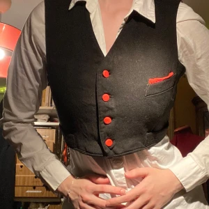 Croppad svart väst med röda detaljer - Snygg croppad svart väst med röda knappar och en röd detalj vid bröstfickan. Handsydd! Köpte den i Berlin på loppis men använder aldrig. Lite staining vid kragen. 100% bomull. Fickorna är dekoration. 