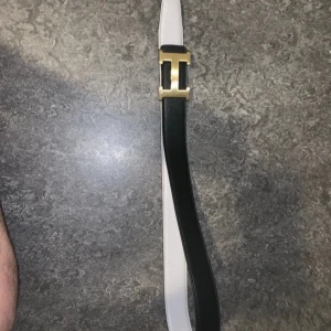 Svart och vitt vändbart bälte med guldfärgat spänne - Snyggt vändbart bälte i svart och vitt med ett stilrent guldfärgat H-spänne. Perfekt för att matcha olika outfits. Tillverkat i skinn för en lyxig känsla.