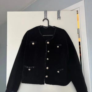 Svart jacka med knappar - Snygg svart jacka med dekorativa knappar framtill. Jackan har en klassisk design med två fickor och en rund halsringning. Perfekt för att ge en elegant touch till din outfit.