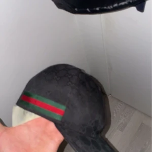 Svart keps med mönster och ränder - Snygg svart keps med diskret mönster och ikoniska ränder i grönt och rött på sidan. Justerbar rem baktill för perfekt passform. Perfekt för en stilren look.