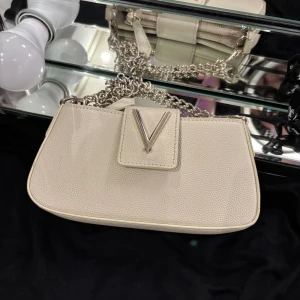 Valentino Handväska - Säljer denna super fina beiga Valentino väskan, då den inte längre kommer till användning. Den är en mindre vässka med dragkedja och innerfickor, samt en kortare kedja. Det hängde kedjor från Valentino märket när jag köpte den, men valde att ta bort de helt, då väskan blir mer stilren så tycker jag själv.