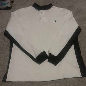 Snygg vit piké, Polo Ralph lauren - Säljer en klassisk vit pikétröja från Ralph Lauren med svart krage och knappslå. Tröjan har en broderad logotyp på bröstet och är perfekt för en stilren look. Passar bra till både jeans och chinos. Tröjan har någon liten fläck på framsidan (se på bilder) skulle tro att det går bort i tvätten. Hör av er om ni har några frågor och priset är såklart inte hugget i sten