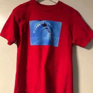 Röd t-shirt från Supreme  - Snygg röd t-shirt från Supreme med ett coolt blått tryck på framsidan. Tillverkad i 100% bomull och har en klassisk passform. Perfekt för dig som vill ha en stilren look med en touch av streetwear.