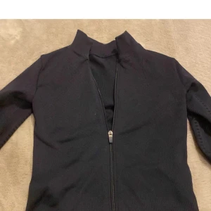 Svart tröja med dragkedja - Säljer en svart tröja med dragkedja framtill. Tröjan är tillverkad av 90% polyamid och 10% elastan, vilket ger en stretchig och bekväm passform. Perfekt för kyligare dagar.