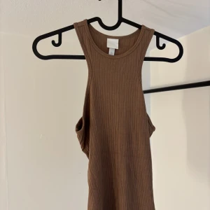 Brun ribbad linnetopp - Säljer en snygg brun ribbad linnetopp. Toppen är ärmlös och har en tight passform, perfekt för varma dagar. Den är gjord i ett mjukt och stretchigt material som sitter skönt mot kroppen.