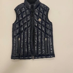 Marinblå Moncler väst - Snygg svart quiltad dunväst från Moncler med dragkedja och två fickor framtill. Västen har en klassisk design med Moncler-logga på bröstet. Perfekt för kyligare dagar. Mycket bra skick endast dragkedjan som är funktion mer går säkert att fixa, skriv vid minsta fråga eller fundering💯👍