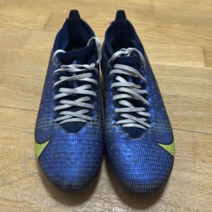 Blå fotbollsskor från Nike - Snygga blå fotbollsskor från Nike med gula detaljer och snörning. Skorna har en modern design med en slitstark sula som ger bra grepp på planen. Perfekta för fotbollsträning och matcher.